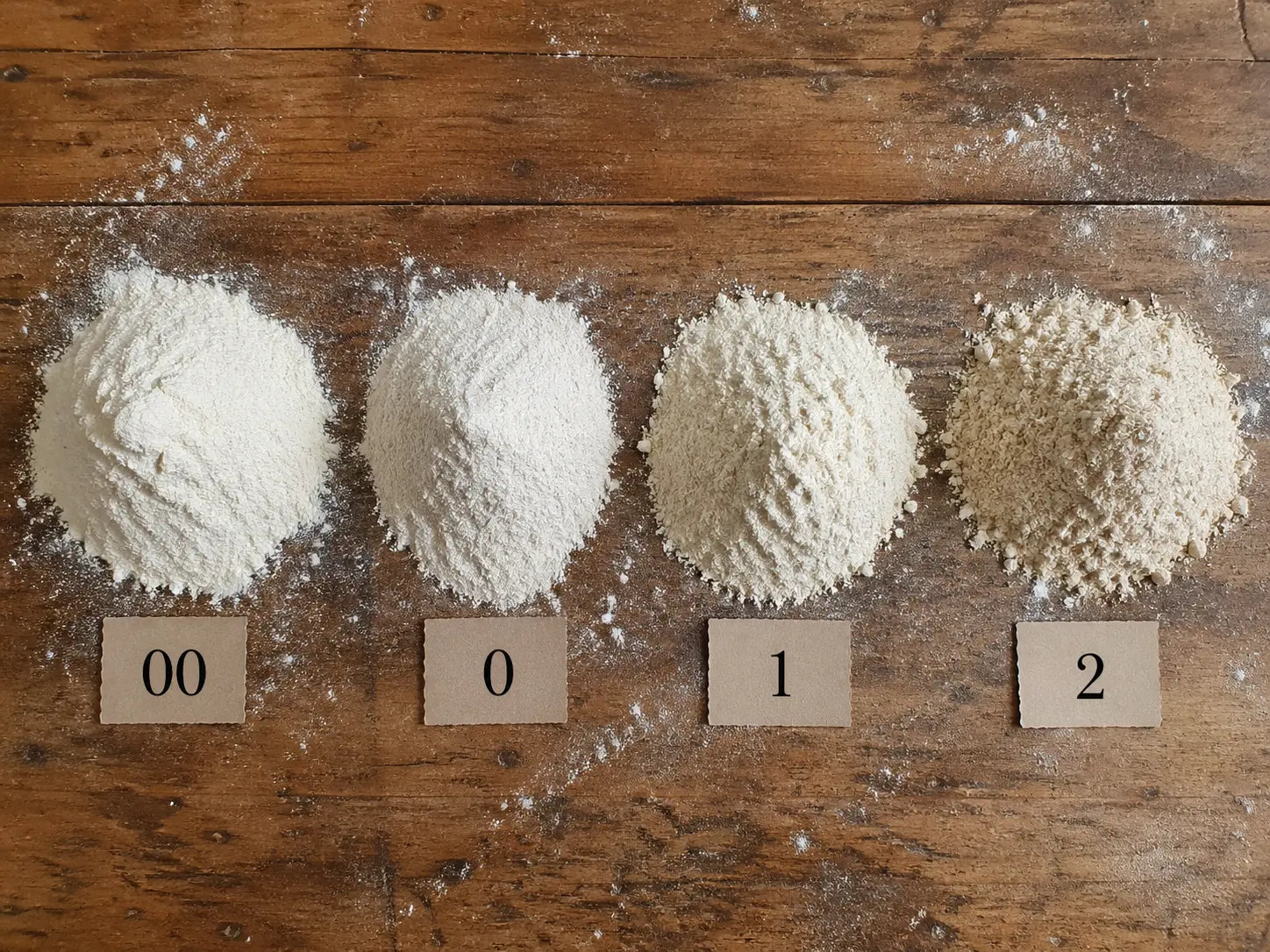 Quattro mucchi di farina 00, 0, 1 e 2 disposti su un tavolo di legno infarinato, ciascuno identificato da un’etichetta numerata.