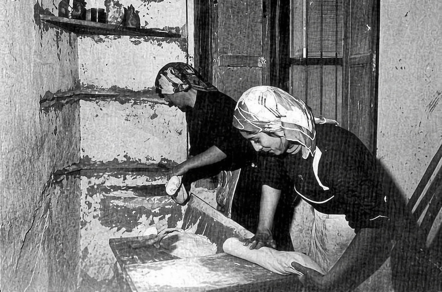 Due donne impastano e lavorano il pane di Matera in un ambiente domestico tradizionale dei Sassi di Matera, in una scena storica in bianco e nero.
